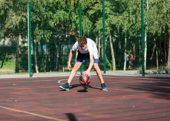 Obraz premium Cute teenager plays basketball at city playground. A boy holds basketball ball in his hands outside. Active life, hobby, sports for children 