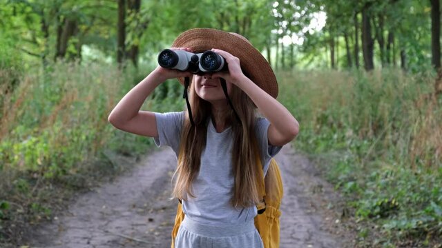 Little girl tourist in hat with backpack looking through binoculars in the forest with a surprised face, camping in the woods. Happy child outdoors. Travel concept