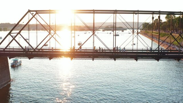 People walk across steel bridge over river. Backlight by sunset. Community fun, festive activity.