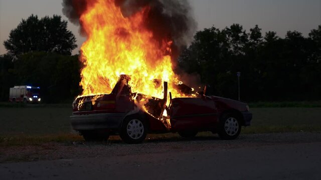 Burning car in the evening hours, firefighters approaching in the background, long shot from the right rear - Slow Motion