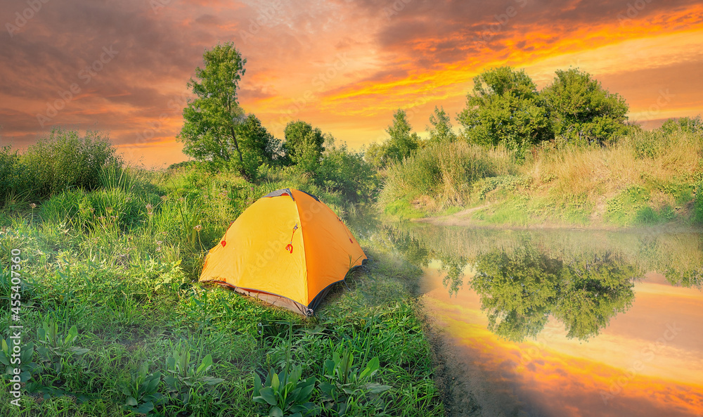 Wall mural orange tent on river bank in green grass under dramatic sky at sunset