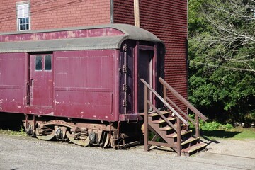 Fototapeta na wymiar Vintage antique railway car with wooden stairs next to a weathered red building with evergreen forest beyond