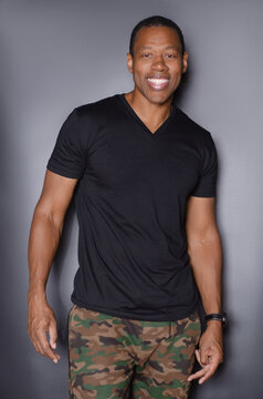 Portrait of  smiling African American man in plain black t-shirt and camouflage pants standing in front of gray background