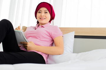 Asian woman on pink shirt, black trousers, cover head by red scarf as breast cancer patient, up knee to take sad note on paper while relaxing on house bed beside alarm clock, photo frame