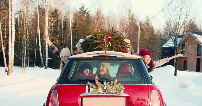 CHRISTMAS FESTIVE MOOD. Three young happy women wave at camera from fancy red car on sunny snowy winter road slow motion