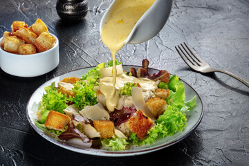 Chicken Caesar salad with the classic dressing being poured, croutons, and pepper, on a black...