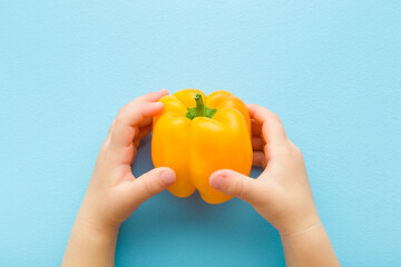 Baby hands holding yellow big sweet paprika on light blue table background. Pastel color. Fresh vegetable. Closeup. Point of view shot. Top down view.