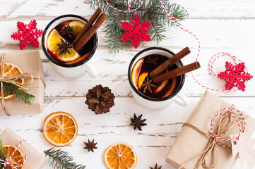 Mulled wine in white enameled mugs with spices and citrus fruit on wooden table with fur tree branches and christmas presents.