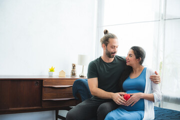 Obraz premium Beautiful young pregnant woman sitting on bed with loving and caring husband trying to connect with baby by touching and feeling stomach at home