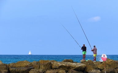     Two adult men are fishing with fishing rods in the sea from the coastal rocks. A sailing yacht is visible on the horizon. 