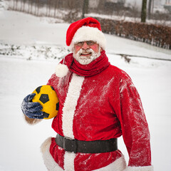 Happy cheerful Santa Claus in a traditional red costume standing on a snowy street with a yellow ball.