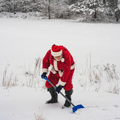 Santa Claus in a traditional red costume with a shovel removes snow on a snowy field.