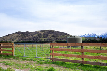 A wooden farm fence with a gate establishes the farm boundary from the country road in Canterbury, New Zealand. A water tank can be seen in the field. - obrazy, fototapety, plakaty