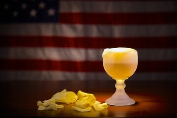 Ice cold schooner glass with beer in front of an American flag with some potato chips on the side.