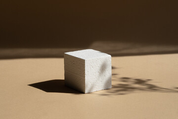 Geometric 3d foam podium for product demonstrations. White cube, in rays of sunlight, casts hard shadow on brown background