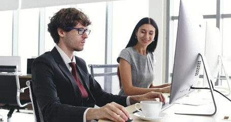 Businessman hipster looks working on desktop computer with serious and focus stressed face near businesswoman colleague, she noticed that and tell him relax and drink coffee and they smile together.