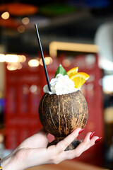 Cocktail made of cracked coconut against blurred red background. Delicious alcohol drink in a coconut shell