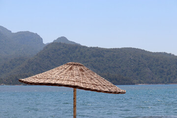 Wicker parasol on blue sea and green mountains background. Beach umbrella, vacation concept