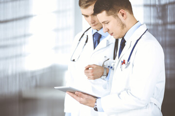 Two doctors with stethoscopes discuss medical exam resoults standing at hospital office. Physician and his colleague use a computer tablet for checking medication names records. Perfect medical