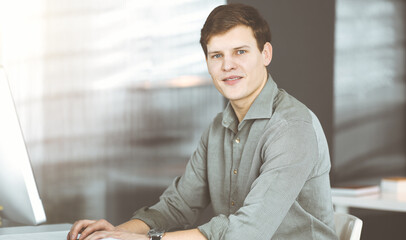 Young businessman working on his computer sitting at the desk in office. Headshot portrait of a man