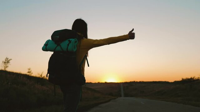The concept of traveling and hitchhiking. A young female traveler hitchhiking and stopping car with thumbs up on road. A female hitchhiker by the road during vacation trip. Summer vacation adventure.