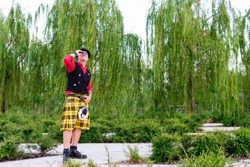 funny and unusual 50-year-old man, dressed as a Scotsman in a kilt and vest, explores a city park and wonders where to find him - obrazy, fototapety, plakaty