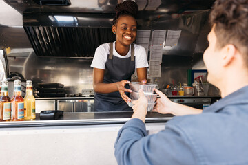 Young woman in apron working at food truck. Small business owner giving takeaway packaged food to...