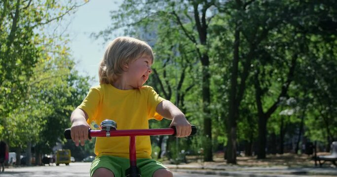 Happy blond little boy wearing yellow t-shirt rides red tricycle along road in green park on sunny summer day close view