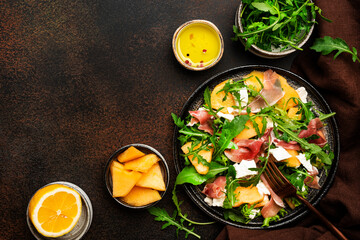 Salad with melon, jamon, goat cheese and arugula on brown background, top view, copy space