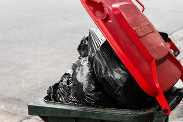 Black plastic bags over flowing modern trash bin with red lid.