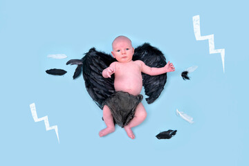 A newborn baby boy with dark demon wings on a blue studio background, copy space. An infant caucasian child in a diaper aged one month