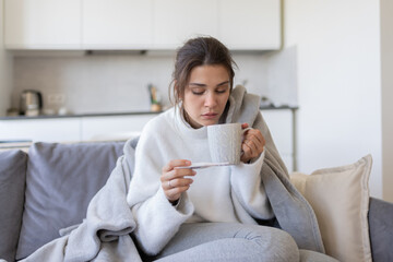 Sick  girl with fever catching a cold, measuring temperature with thermometer hold cup of hot tea or medicine sitting on couch at home. Ill woman covered with blanket got grippe, flu concept.