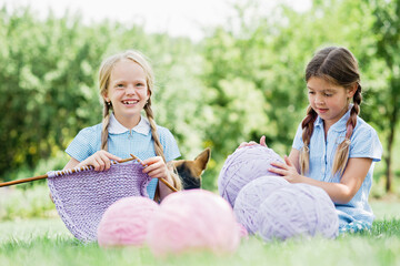 Two girls sitting on green grass and knitting sweater with needles on summer day.