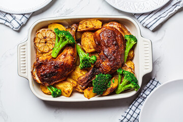 Baked chicken legs with potatoes, broccoli and garlic in baking dish, white marble background. Healthy festive food concept.