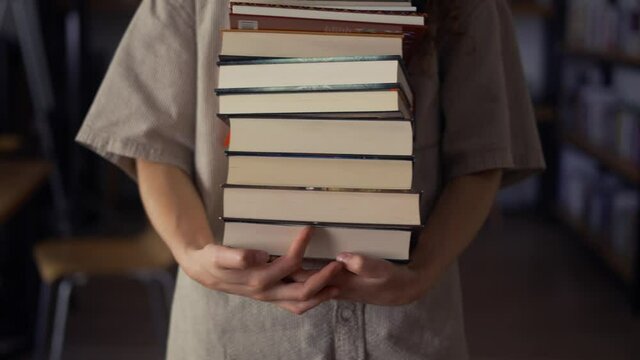 Unrecognizable student walks with plenty of books in the library, preparing for exams