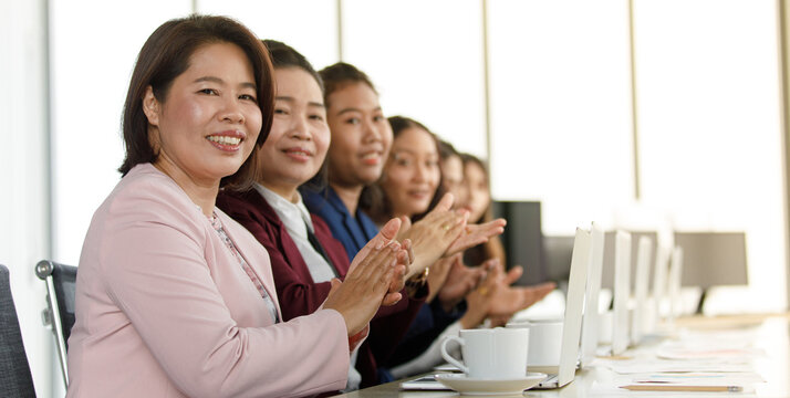 Group of businesswomen in suit sitting at working desk and clapping hands for appreciating to someone in modern office company. Concept of good circumstance in company with friends and colleagues