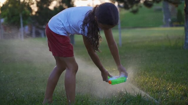 Girl draws water from the lawn sprinkler into the water gun. Happy kid play with a stream of water from a lawn sprinkler