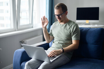 Cheerful young man using laptop sitting on the comfortable couch, guy is waving hello into webcam, smiles and greeting. Video call, video conference, online meeting on the distance