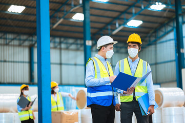 Team engineers and foreman wear a mask, hard hat, and vest. Standing, consult discuss industrial production management through data files with in mask factory. Employees working in the background.