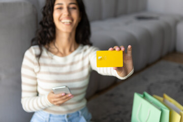 Happy young woman showing credit card at camera, holding smartphone, shopping in online store from home, selective focus