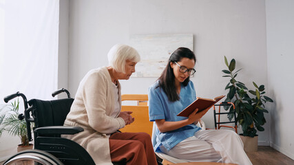 brunette geriatrician nurse reading book to aged woman in wheelchair