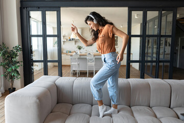 Positive young woman with headset dancing on couch while listening to music at home, full length