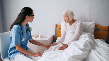 geriatric nurse in eyeglasses talking with aged woman in bed