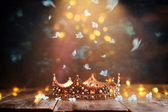 low key image of beautiful queen or king crown over wooden old table and falling flowers. fantasy medieval period