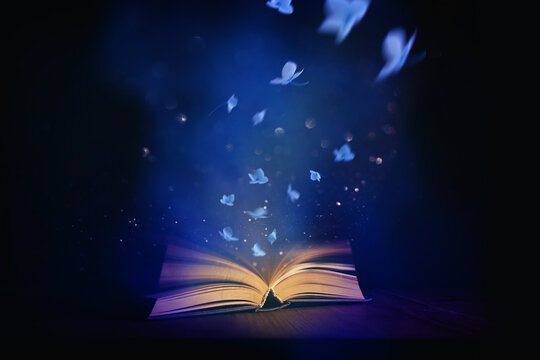 Magical image of open antique book over wooden table with glitter lights and flowers flying from it