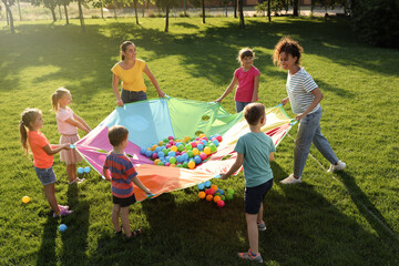 Group of children and teachers playing with rainbow playground parachute on green grass. Summer...