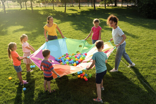 Group of children and teachers playing with rainbow playground parachute on green grass. Summer camp activity