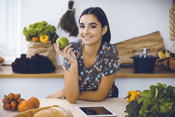 An attractive young dark-haired woman holding green apple and choosing the recipe for a delicious meal while smiling in the kitchen. Tablet pc is the best cookbook