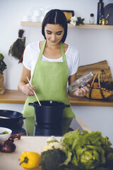 An attractive young dark-haired woman preparing soup by new keto recipe while standing and smiling in the kitchen. Cooking and householding concepts
