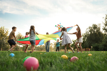 Group of children and teachers playing with rainbow playground parachute on green grass, low angle view. Summer camp activity - obrazy, fototapety, plakaty